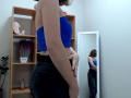 Steisii stands in a room, wearing a blue strapless top and black pants, with her back to a mirror reflecting her ass and torso. She has fair skin and brown hair.