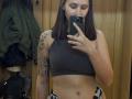 HotCaramelsTrio with brown hair, wearing a gray sports bra, showing a tattoo on her left arm, holding a phone, reflecting in a mirror, showing her tits and belly.