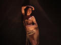 TriciaBarnes with bare tits, tied in rope, wearing a golden cloth, standing in dark, with arm above head, and leg slightly raised, smiling, looking right, soft lighting.