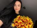 Olieri with long black hair, blue eyes, and a smile, holding a bouquet of yellow tulips with red centers, wearing a black shirt.