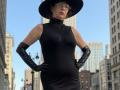 MaitresseElsa stands in a city, wearing a sheer black dress, black gloves, and a wide-brimmed hat, with hands on hips, and glasses, showing her tits and ass.