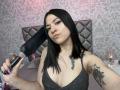 GoddessLuxliv with black hair, nose ring, and tattoos, holding a black hairdryer, wearing a glittery black dress, showing her tits, in a bedroom with pink and gray decor.