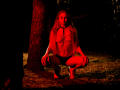 MiaMeowa crouches in dark red light, wearing chains across her bare tits, black shorts, and heels, with long blonde hair, in a forest at night.
