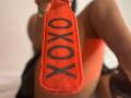 LilithRussoo holding a bright orange, textured "OXO" strap in front of her, with her large, bare tits slightly visible in the blurred background.