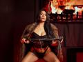 AfroditaDoir with long black hair, wearing a fur coat, black lace lingerie, and holding a whip, sitting with her tits and pussy exposed, in front of a fire.
