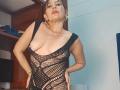 NayaSex com seios grandes e expostos, vestindo um vestido de malha preta transparente, em pé com as mãos nos quadris, batom vermelho, brincos de argola dourados e uma expressão confiante e sexy..