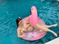 MaddyCarina with tan skin, dark hair, and pink flower in her hair, lying in a pink inflatable unicorn, wearing a silver bikini, exposing her tits, in a blue pool.