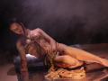 TriciaBarnes lying on a marble slab, wearing a rope harness that barely covers her tits and pussy, with a rope in her hand, in a smoky, dimly-lit setting.
