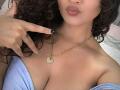 CanelaLebrad with curly dark hair, pouty lips, wearing a gold necklace with a heart-shaped diamond, pointing at it with black-manicured nails, in a blue off-shoulder top.