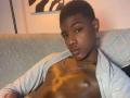 DrakeWit, black dude, short curly hair, shiny brown skin, no shirt, gray cardigan, chain necklace, sitting on patterned couch, looking at camera, sexy, cute, relaxed.