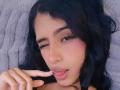 LilithDesire, with dark wavy hair, one eye closed, licking her finger, has pink lipstick, dark eyeliner, and a cute, sexy expression. Her lips are slightly parted.