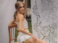 EvaDaly with small tits and a frilly white top and shorts, sitting on a wooden chair, looking over her shoulder, lace curtain to the right, outdoor setting.