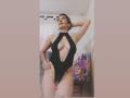 ChanelJolie in a black cut-out one-piece, showing her tits and pussy, standing with one hand behind her head, looking up. Tattoo on right thigh. Indoor setting.