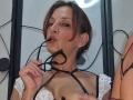CassandraMichelli with brown hair, wearing white lace top, holding black glasses in her mouth, showing her tits, black strap choker, and small earrings, in a selfie.