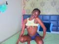 KharlaQueen is crouching on a green bed, topless with small tits, wearing blue panties, smiling at the camera. She has dark skin and braided hair.