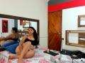 AlisonHank with long black hair, black tank top, and white panties, sitting on a bed with blue pillow, showing her ass, with a mirror reflecting her back.