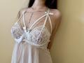 Tisia wearing a white lace babydoll with bows, her tits covered in delicate lace, chains hanging from her neck, and a sheer lower part, against a beige wall.
