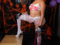 JeimyCampos in white lingerie with red stains, sheer white garter, white thigh-highs, black skeleton gloves, holding red and black straps, one leg on a box, showing her tits.