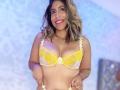 AuroraBenson with medium brown skin, wavy blonde hair, smiling, wearing a yellow floral bra, showing her tits, holding the bottom of her bra, red nail polish.