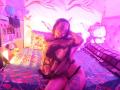 MilaThorne with glasses, wearing a sheer floral top, black thong, and tattoos, sits on a colorful bed, pink lighting, hand in hair, looking at camera.