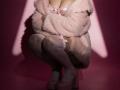 Destinis crouches with pale pink fur coat, white lace bra, and thigh-high stockings, wearing pink platform heels. Boobs partially covered, arms crossed, shadowed in pink light.
