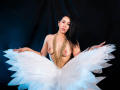 Katty Rivera, avec ses cheveux noirs, son collier en or et sa jupe blanche ornée de plumes, est nue, montrant ses seins, les bras tendus, sur un fond sombre..