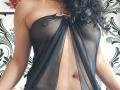 SexyMirra with curly black hair, wearing a sheer black top that reveals her small, perky tits and her bare, flat, brown-skinned belly. Top has a gold ring in center.