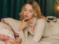 KylaHemeke with blonde hair, red lipstick, and pearl necklace, lying on a bed, wearing a white long-sleeve top, touching her chin, looking down, with pink and floral pillows.