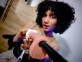 CinthiaWalker with curly black hair, holding a pink dildo in front of her, looking at the camera, wearing a grey top, wooden floor, and a black chair.