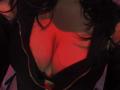 KiraBerry's large, red-lit tits are visible through a black, zipped-up top with a deep cleavage, showing black bra straps. Her dark, wavy hair partially covers her face.