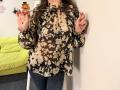 Tilliana, com uma blusa floral marrom, segurando fitas com um prendedor de abóbora, unhas rosa, jeans azuis e cabelos castanhos longos, em pé em frente a uma parede branca..