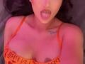 ItsJessieBabe, mit herausgesteckter Zunge, ein orangefarbener BH, der ihre Brüste zeigt, ein kleines Tattoo über ihrem Dekolleté und eine rosa-rote Beleuchtung auf ihrer hellen Haut..