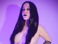 KendraSimons with long black hair, purple lighting, and dark lipstick, topless, showing her tits, wearing fishnet arm sleeves, looking at the camera with a slight smile.