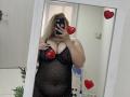 DiLaria, avec ses cheveux blonds, portant une robe noire transparente avec des paillettes, montrant ses seins et un tatouage au-dessus de sa poitrine, prend un selfie devant un miroir avec des autocollants en forme de cœur rouge..