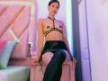 MelisaDaltonn with bare tits, black strappy harness, black latex skirt, and thigh-high boots, sitting on pink bed, looking at camera, dark hair, light skin, confident expression.