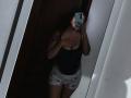 AntoniaBrunni in mirror selfie, wearing black tank top covering her tits, grey shorts with blue hearts, dark hair, holding phone with blue case, shadowy room.
