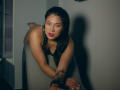LindaJimenex with dark hair, red lipstick, and black leather collar, crouching on gray furniture, showing tattoos on her arm, looking seductively at the camera with handcuffs in front.