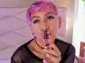 DannyRouds with pink hair, black mesh top, red choker, holding a purple dildo, looking up with open mouth, wearing black ear gauges, in a brightly lit room.