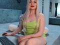 CoquineElise with long platinum blonde hair, red lipstick, green tank top, sitting on bed, legs together, arms resting on keyboard, looking at camera, showing small tattoos on arms.