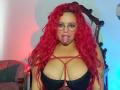 Estefy69 with red curly hair, glasses, tongue out, black strappy top, large tits, and red criss-cross straps. She's looking at the camera, standing in front of a dark mirror.