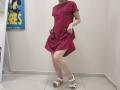 JoannaSia wearing a red dress, lifting it to show her bare ass, standing in white shoes, pale skin, and a white wall background. Her face is not visible.