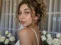 Sanella has curly blonde hair in a ponytail, blue eyes, and pink lips, wearing a white top, with white roses in the foreground, and gold earrings.