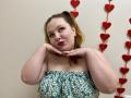 Ailia, a plus-sized woman with fair skin and red hair, poses with hands under chin, wearing a strapless floral top, against a wall with red heart decorations. Seductive expression.