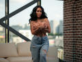 NyraNoble, topless with hands covering her tits, has tattoos on her arms and chest, wears ripped jeans, standing in a modern room with brick walls and city view.