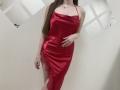 Nesie wears a shiny red satin dress with a lace side, showing her ass and a leg. Long brown hair, thin necklace, hand on her hip.
