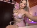 EliseMonet, avec ses longs cheveux blonds platine, porte de la lingerie rose transparente avec des broderies de papillons, montrant ses seins, assise sur une chaise, tenant sa poitrine, et souriant à la caméra..