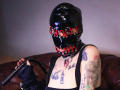 AmySpider wearing a shiny black latex mask with red eyelets, holding a black leather whip, tattooed chest and arms, black dress, sitting on a brown leather couch.