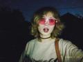 EmelyDilsaver with wavy brown hair, wearing pink sunglasses, a black choker, and a gray shirt, has her mouth open in a surprised or shocked expression. 2026.7.12 timestamp.