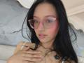 SophieDelaney with pink glasses, black hair, pink blush, nose ring, and white tank top, hand on chest, small black tattoo on right shoulder, pink lips, and subtle smile.