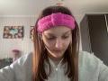 AdrianneEllinger with pink fuzzy headband, gray hoodie, brown hair, and downcast eyes, wearing white earbuds. Her ass is not visible. She has a small beauty mark on her cheek.